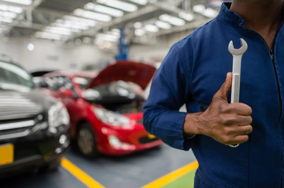 Auto mechanic in a blue uniform holding a wrench inside a busy car repair shop with vehicles in the background, representing professional car servicing and repairs, lemonmyvehicle