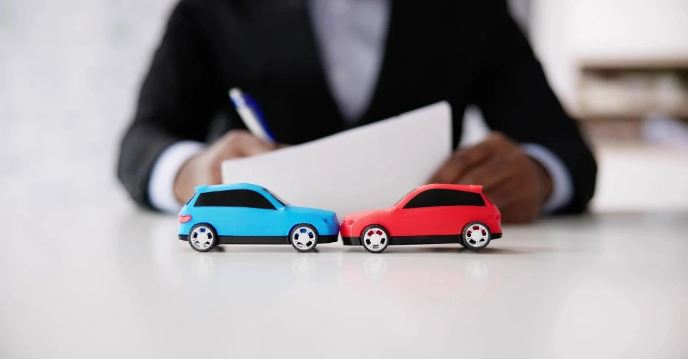 Blue and red toy cars staged in a crash with a businessperson reviewing paperwork in the background, representing car accident claims or insurance disputes, lemonmyvehicle