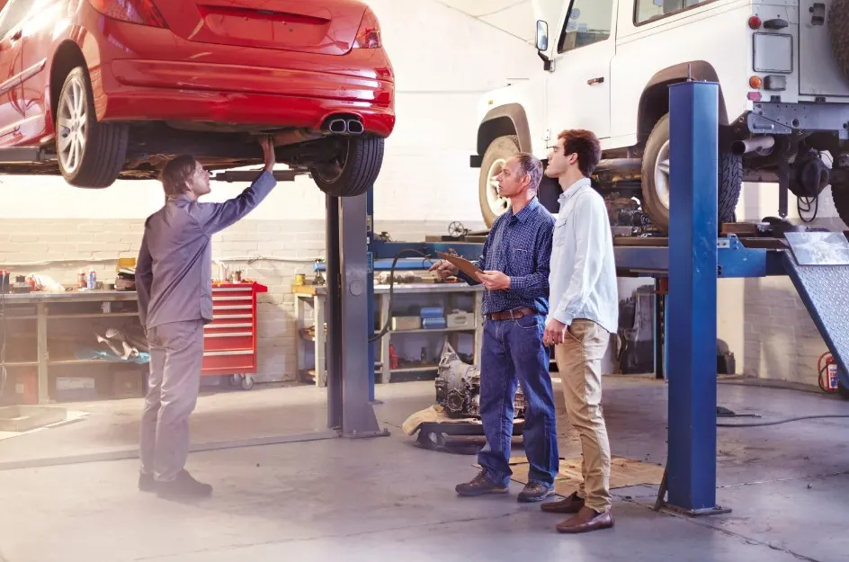 Mechanic inspecting a red car lifted on a hydraulic platform while two customers observe in an auto repair shop, symbolizing vehicle maintenance or pre-purchase inspection, lemonmyvehicle