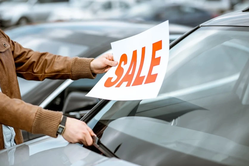 Person placing a red 'SALE' sign on a car windshield at a dealership or used car lot, indicating a vehicle for sale, lemonmyvehicle