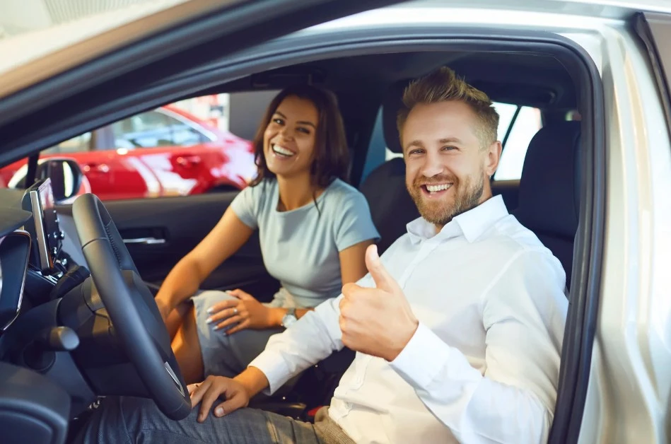Smiling couple sitting inside a new car at a dealership, with the man giving a thumbs-up, symbolizing a successful vehicle purchase or leasing experience, lemonmyvehicle