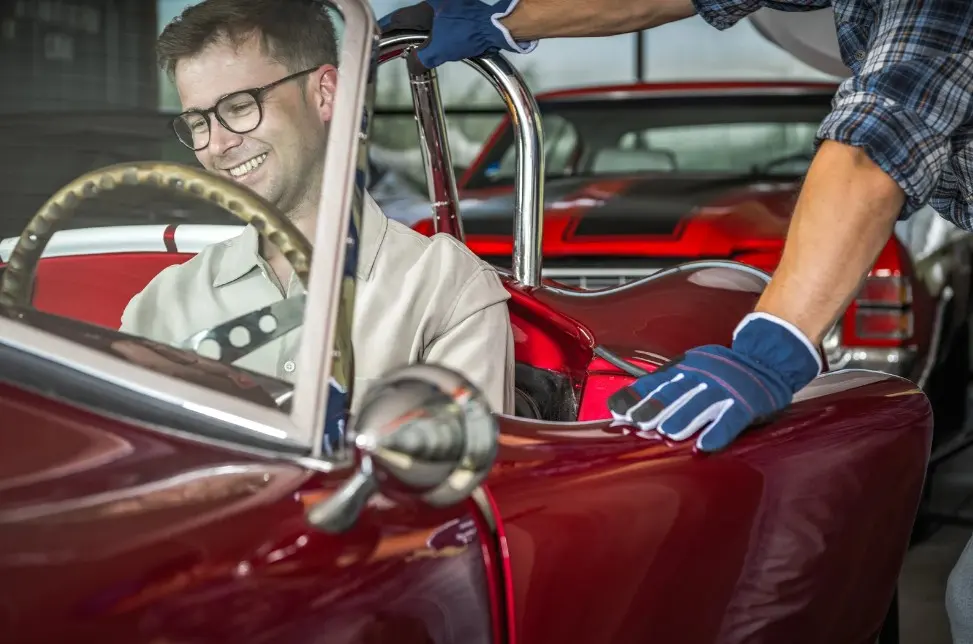 Smiling man sitting in a vintage red convertible while a gloved technician assists, symbolizing a successful classic car purchase or restoration, lemonmyvehicle