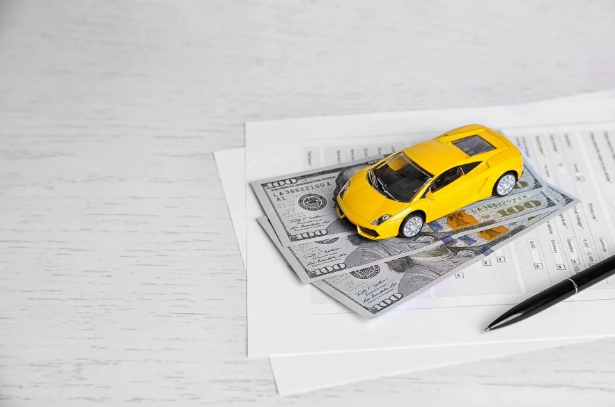 Yellow toy car placed on U.S. hundred-dollar bills and financial documents, representing car loans, auto financing, or vehicle investment, lemonmyvehicle