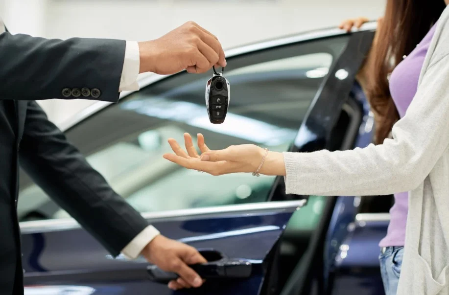A hand holding car keys with a white car in the background, representing vehicle ownership and the potential for a new car to be a lemon.