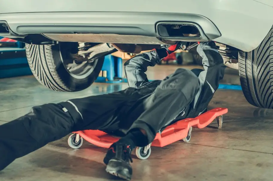 A mechanic working underneath a car on a creeper, illustrating the diagnostic and repair processes for defective vehicles.