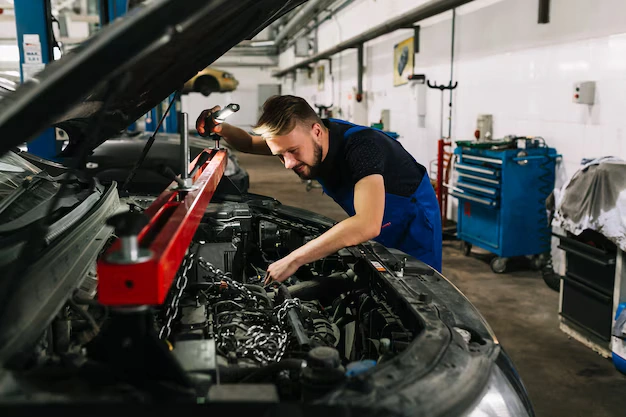 A male auto mechanic working on a car engine in a garage, using a work light to illuminate the area.