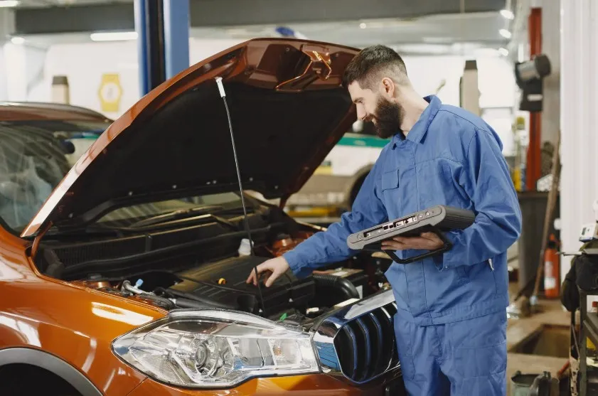 A mechanic in a blue uniform working on an orange car's engine with a diagnostic tablet, indicating vehicle inspection and repairs relevant to Lemon Law cases.