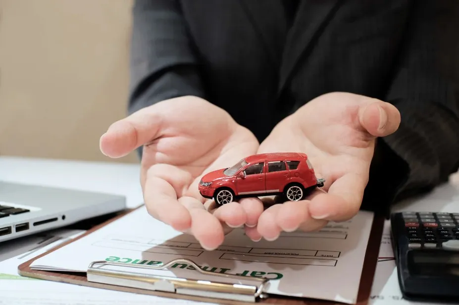 Person holding a small red car over car insurance and warranty documents, symbolizing Lemon Law vehicle protection