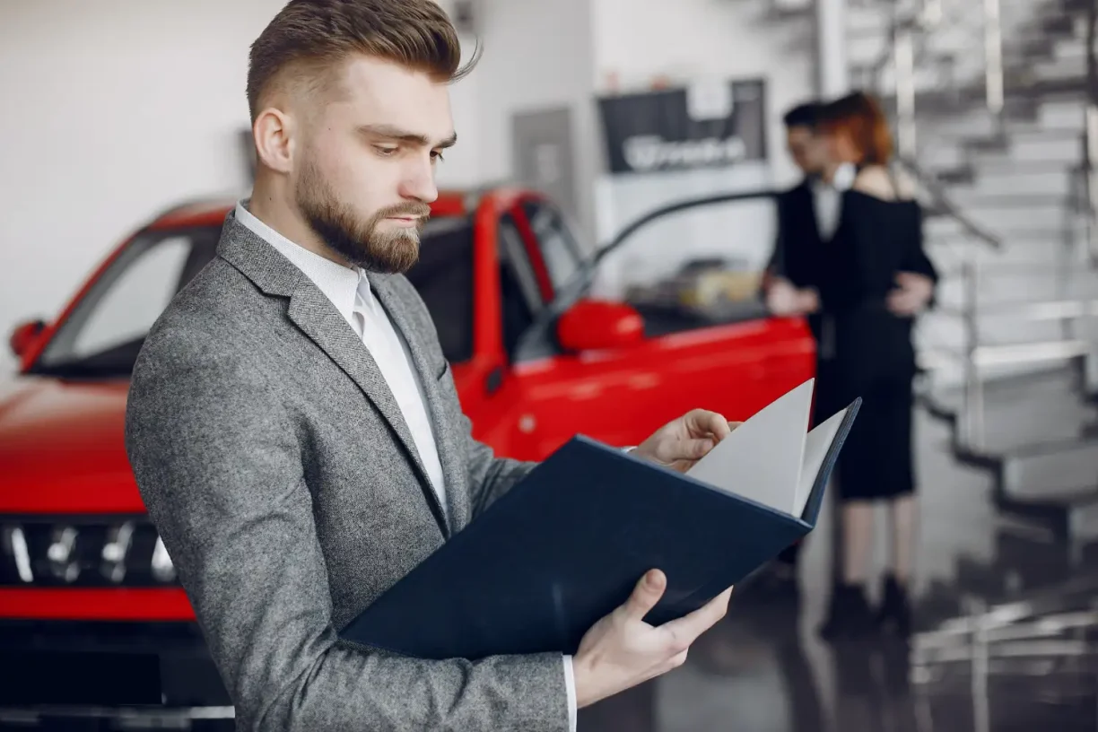 car lawyer in a suit reviews a brochure or documents in a showroom, with a red car and other customers in the background.